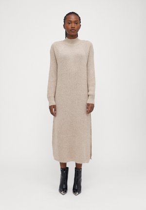 Beiges, gestricktes Midikleid mit hohem Kragen und gerippten Ärmeln, mit seitlichen Schlitzen. Trägt dazu schwarze Ankle Boots mit spitzen Zehen.