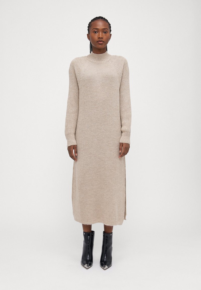 Μπεζ πλεκτό midi φόρεμα με ψηλό λαιμό και ελαστικά μανίκια, που διαθέτει πλαϊνές σχισμές. Φορεμένο με μαύρες μποτες στον αστράγαλο με μυτερούς τάρσους.