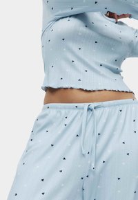 Lichtblauw pyjama set gemaakt van zachte stof. Het bovenstuk heeft een geribbelde textuur met kleine hartjespatronen; de broek heeft een elastische tailleband en een trekkoord.
