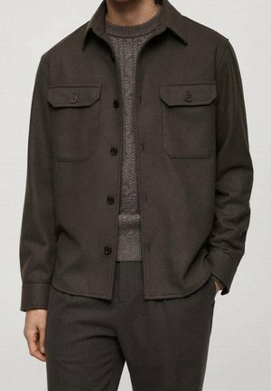 Homme portant une veste marron foncé boutonnée avec poches à rabat par-dessus un pull gris texturé et un pantalon foncé, main dans la poche sur un fond clair.