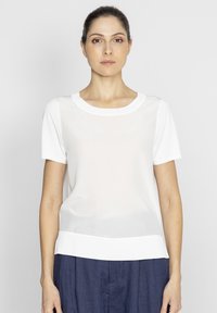 Biała bluzka z krótkim rękawem i okrągłym dekoltem, wykonana z przezroczystego materiału. Posiada solidny, biały brzeg, w zestawieniu z luźnymi, granatowymi spodniami.