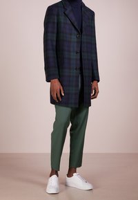 Soprabito a quadri blu navy e verde con rever a incavo, tre bottoni e tasche laterali, abbinato a pantaloni verdi su misura e sneakers bianche.