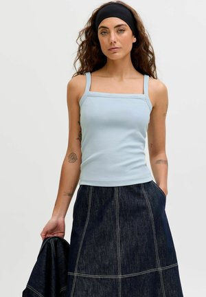 Kvinde med bølget hår og pandebånd iført lyseblå top med firkantet halsudskæring og mørk denimnederdel, der holder en matchende denimjakke i hånden.