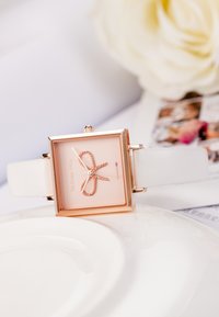 Reloj cuadrado con caja de oro rosa, esfera rosa y diseño de lazo, que cuenta con una correa de cuero blanco y un detallado minimalista.