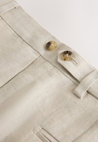 Pantalon en lin beige avec deux boutons de style corne à la taille et une poche latérale visible sur la gauche.