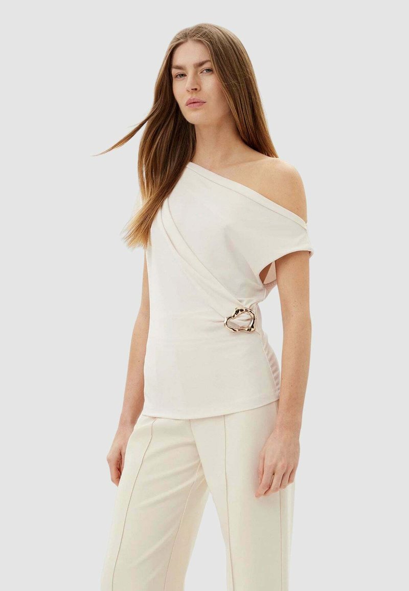 Top color crema con spalle scoperte e tessuto drappeggiato, caratterizzato da un dettaglio in metallo dorato in vita. Abbinato a pantaloni color crema su misura. Tessuto liscio.