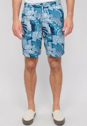 Homme portant un short bleu à motif tropical, des chaussures blanches sans lacets et une chemise blanche, debout devant un fond clair et uni.