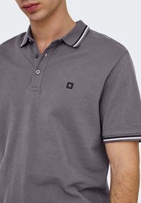 Mand iført en grå polo med hvid- og sortstribet krave og ærmekanter, med et lille sort firkantet logo på brystet.