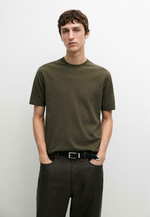 Jonge man met kort bruin haar, die een donkergroene korte mouw t-shirt en een zwarte riem met donkere broek draagt, staand met handen in de zakken.