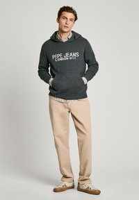 Sudadera gris oscuro con "PEPE JEANS LONDON W11" impreso en blanco, combinada con pantalones beis claros y zapatillas multicolor.