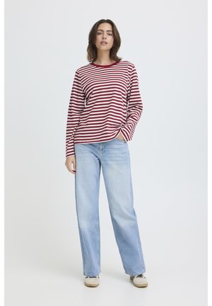 Femeie în picioare, purtând un tricou cu mâneci lungi în dungi roșii și albe, blugi largi de culoare albastru deschis și pantofi bej, pe un fundal simplu.