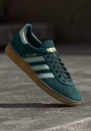 Baskets Adidas Spezial bleu canard foncé avec bandes bleu clair et semelle en gomme flottant au-dessus d'une surface en béton.