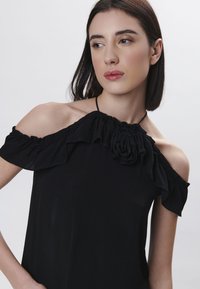 Top nero con spalle scoperte, caratterizzato da dettagli a balze e un fiore decorativo sul collo, realizzato in un tessuto leggero e liscio.