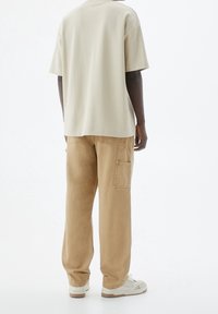 Osoba ubrana w beżowy, oversize'owy t-shirt, luźne beżowe spodnie cargo z bocznymi kieszeniami i białe sneakersy, stojąca na tle białej ściany.