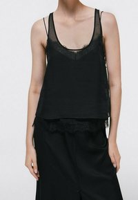 Camisole nera con spalline sottili, caratterizzata da un bordo in pizzo sul colletto e sull'orlo. Realizzata in un tessuto leggero e semi-trasparente, dal fit ampio.