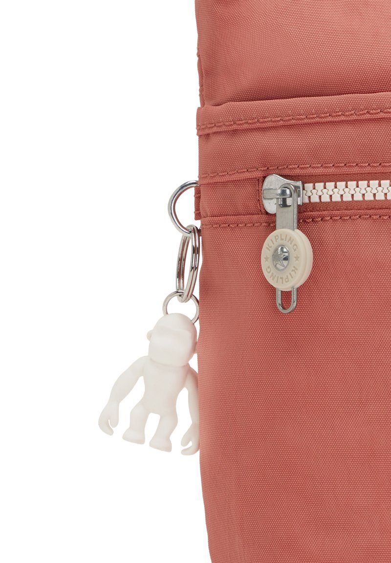 Primer plano de una cremallera de una bolsa de tela color rosa empolvado con un tirador redondo blanco de la marca Kipling y un pequeño llavero en forma de mono blanco adjunto.