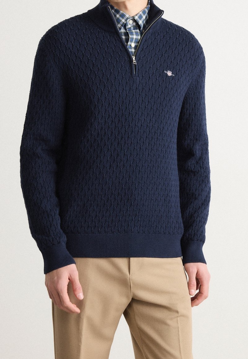 Homme portant un pull texturé bleu marine à demi-fermeture éclair sur une chemise à carreaux bleus avec un pantalon beige, se tenant devant un fond clair uni.