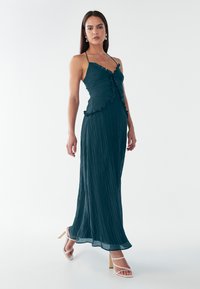 Vestito maxi plissettato in teal con bordi arricciati, spalline sottili e corpino aderente. Abbinato a sandali bianchi con tacco. Tessuto morbido e fluido.