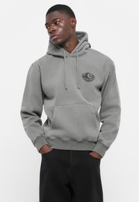 Grijze hoodie met een voorzak, voorzien van een ronde afbeelding op de linkerkant van de borst. De stof lijkt zacht en gestructureerd.