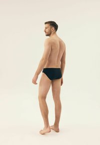 Czarne kąpielówki na męskiej sylwetce lekko odwróconej, z krótkimi włosami, jasną brodą i bosymi stopami na neutralnym tle.