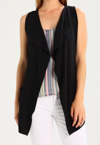 Black sleeveless cardigan with an open front design, featuring a soft texture and draped look, paired with a colorful striped top underneath.