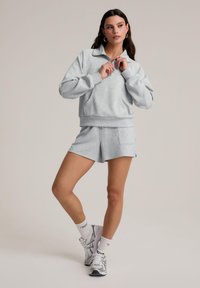 Sweatshirt court gris clair avec un col zippé et un short assorti, présentant une texture douce. Porté avec des chaussettes blanches et des baskets grises.