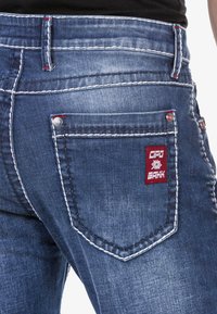 Verblasste blaue Jeans mit weißen Nähten, die ein rotes Logopatch auf der Gesäßtasche und silberne Nieten zur Verstärkung aufweisen.