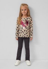 Chemise beige à manches longues avec un motif léopard noir et rose, ornée d'un cœur en sequins. Associée à un pantalon ajusté gris foncé et des baskets blanches.