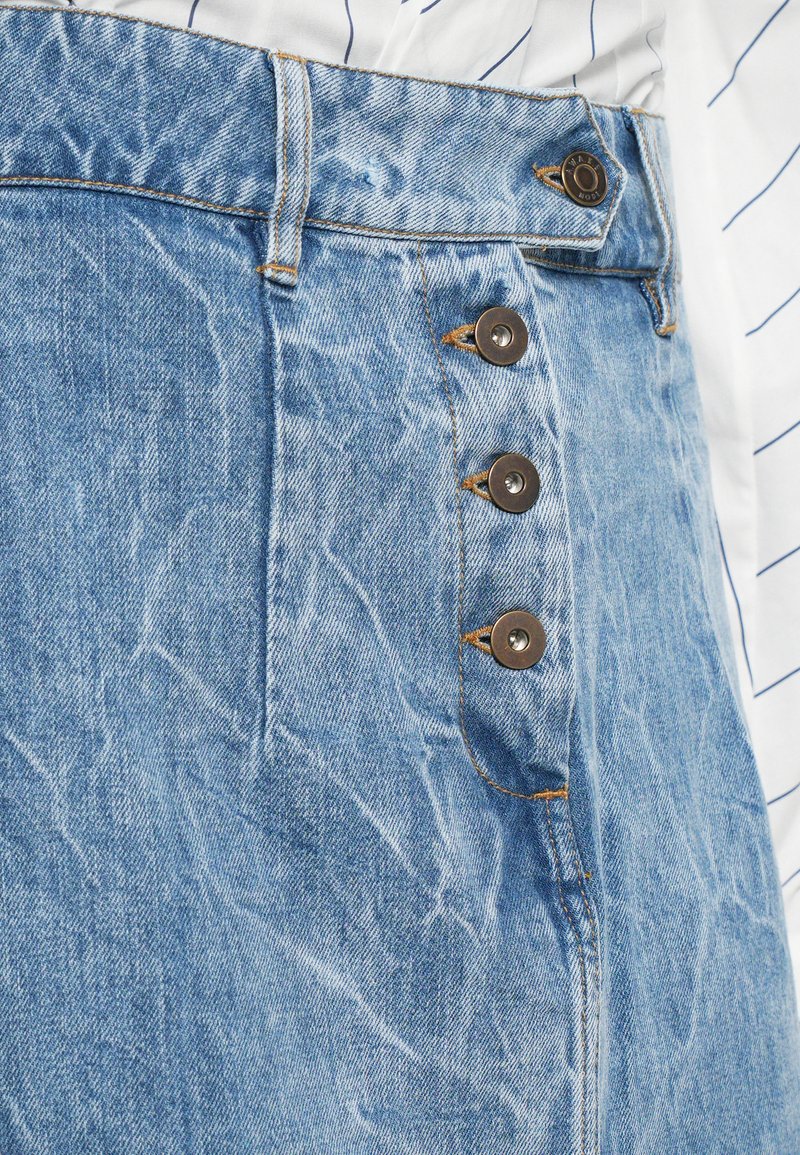 Högmidjad ljusblå denimkjol med tre synliga metallknappar och orange sömnad, bärs med en inskjuten vit skjorta med tunna ränder.