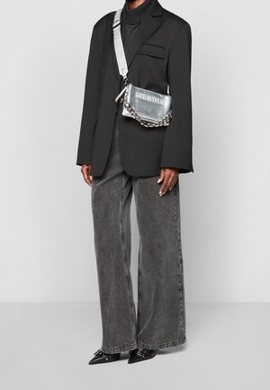 Person iført en sort oversized blazer, grå vidbensjeans, spidse sorte sko og en sølv crossbody-taske med kædedetaljer.