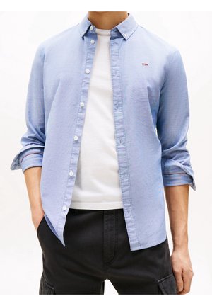 Mann trägt hellblaues Hemd über weißem T-Shirt und schwarze Hose, Hemdsärmel hochgekrempelt, Hände in den Taschen, Torso sichtbar.