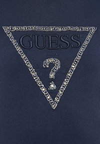 Tejido marino con un logo triangular de GUESS adornado con cuentas, que incluye un signo de interrogación, el texto "U.S.A.", "WASHED" y "JEANS".