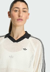 Biele polo tričko z meshu s čiernym golierom a tromi čiernymi pruhmi na rukávoch. Obsahuje malé čierne logo na hrudi.