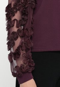 Ilgi piedurkņu tops dziļi bordo krāsā ar caurspīdīgu audumu, piedurkņu daļās dekorēts ar ruskām ziedu akcentiem un solidu ķermeni. Mīksta tekstūra.