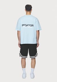Ljusblå bomulls-T-shirt med texten "PEGADOR" och "Next Wave" i svart och vitt. Kombinerad med svarta shorts med vita ränder.