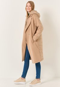 Cappotto lungo beige con cappuccio imbottito, caratterizzato da una vestibilità sartoriale, tasche laterali e una texture liscia. Abbinato a jeans blu e sneakers beige.