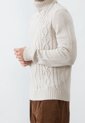 Homme portant un pull à col roulé écru en maille torsadée et un pantalon en velours côtelé marron, vu de profil, les mains jointes.