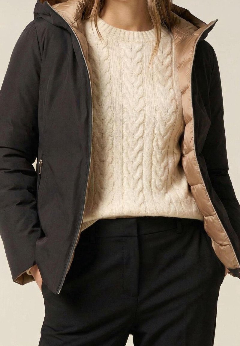 Femme portant une veste noire zippée avec une doublure matelassée beige sur un pull en maille torsadée crème et un pantalon noir, mains dans les poches.