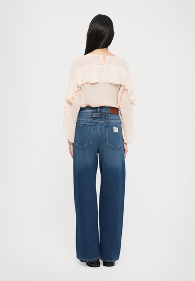 Helle rosa Chiffon-Bluse mit rüschigen Akzenten, kombiniert mit weit geschnittenen blauen Denim-Jeans. Sichtbares Markenlogo auf dem Bund, schlichte Rückengestaltung.