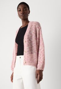 Cardigan lavorato a maglia rosa chiaro con una vestibilità morbida, polsini a coste e chiusura con cinque bottoni, indossato sopra un top nero e pantaloni bianchi.