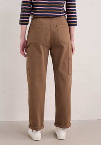 Pantalones cargo marrones con bolsillos traseros, puños enrollados y un ajuste relajado, combinados con una camisa de rayas de manga larga y zapatillas blancas.