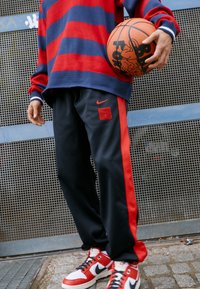 Jersey de punto a rayas rojas y marinas con mangas anchas, combinado con pantalones de chándal negros con rayas rojas en los laterales y un parche de logo, y zapatillas rojas y blancas.