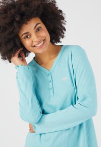 Haut turquoise à manches longues avec texture côtelée, décolleté en V et patte de boutonnage. Présente un petit design de cœur en argent sur la poitrine.