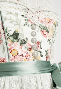 Corset floral avec bordure en dentelle, boutons nacrés et large ruban en satin vert sauge noué au-dessus d’un tissu en dentelle blanche.
