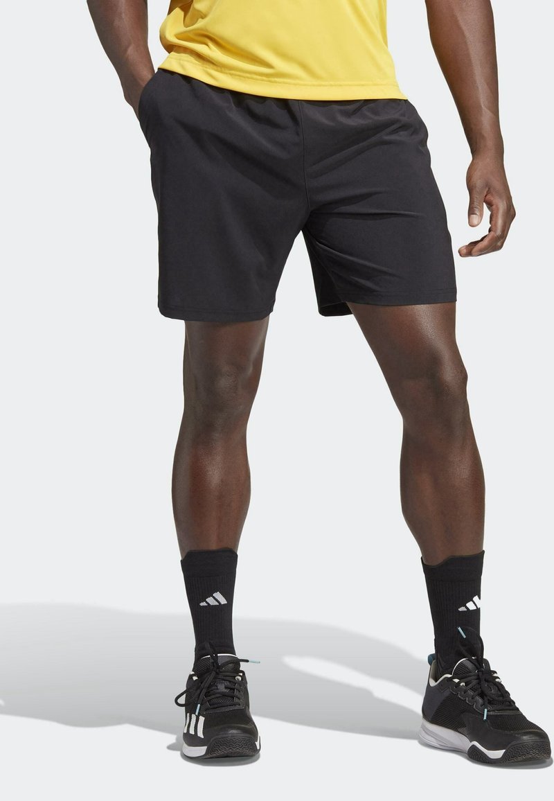 Athlète portant un short de sport noir, des chaussettes Adidas noires et des chaussures de course Adidas noires, debout les jambes écartées sur un fond blanc.