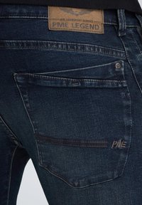 Jeans de mezclilla azul oscuro con una textura suave, que cuentan con una etiqueta de cuero con la inscripción "PME LEGEND" y costuras visibles en el bolsillo trasero.