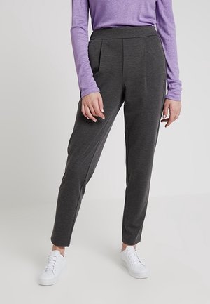 Person iført mørkegrå taperede bukser, hvide sneakers og en langærmet lilla top, der står mod en ensfarvet baggrund.