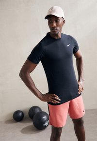 Svart trenings t-skjorte med korte ermer og en liten hvit logo, sammen med lyserosa shorts; bakgrunnen har svarte treningsballer.