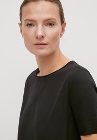 Kobieta o jasnej karnacji i zielonych oczach, ubrana w prostą czarną bluzkę z krótkim rękawem, zwrócona przodem na tle jednolitego jasnego tła.