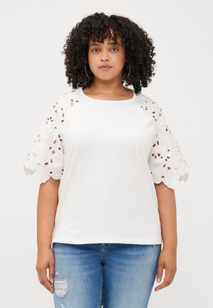 A woman with curly hair wearing a white top featuring floral lace sleeves stands against a plain background, hands relaxed by her sides.